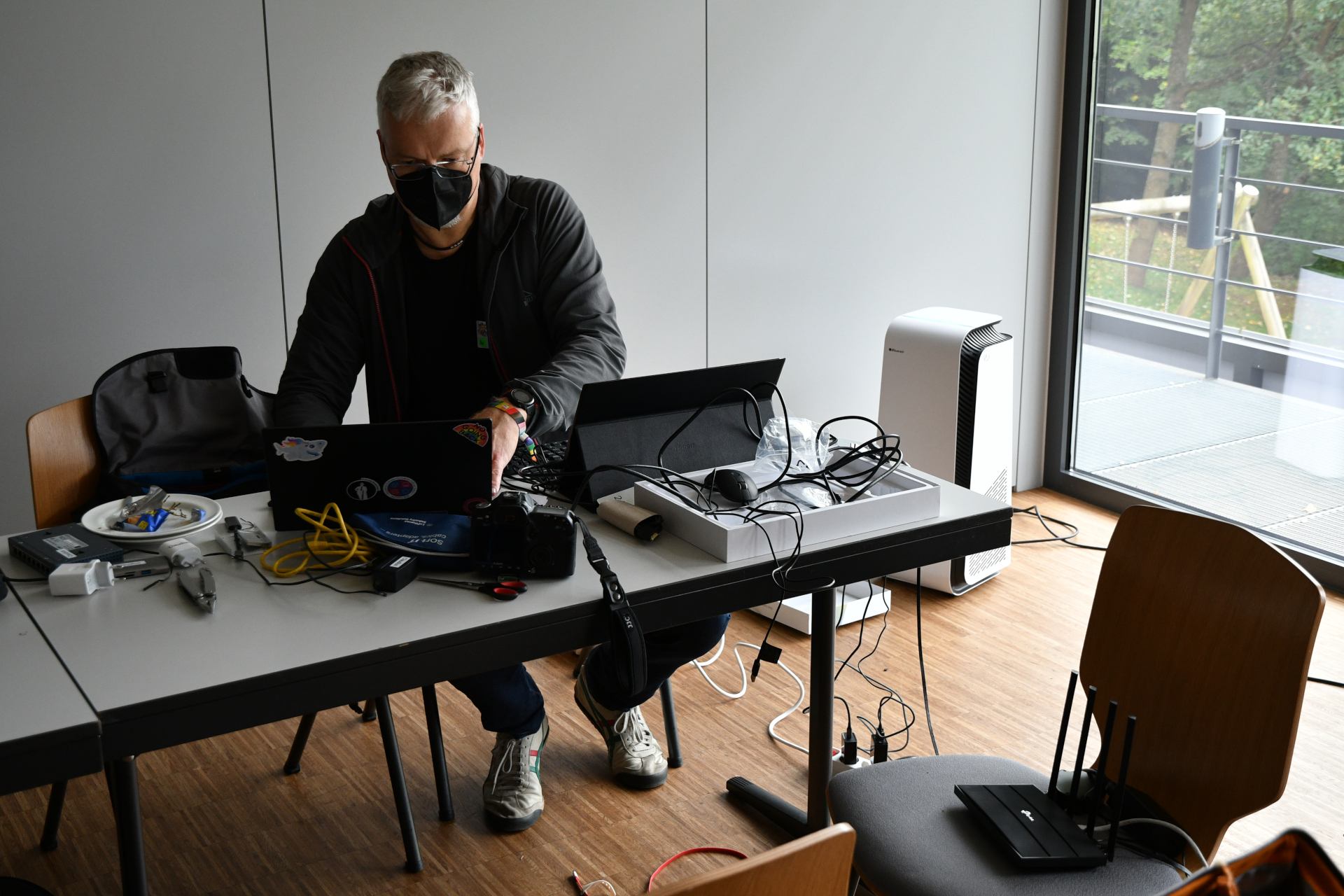 A masked man in dark cloths sits at a table. On the table is a lot of technical equipment like a camera, a laptop, a tablet, some tools, multiple cables for ethernet and power and also a multitool.