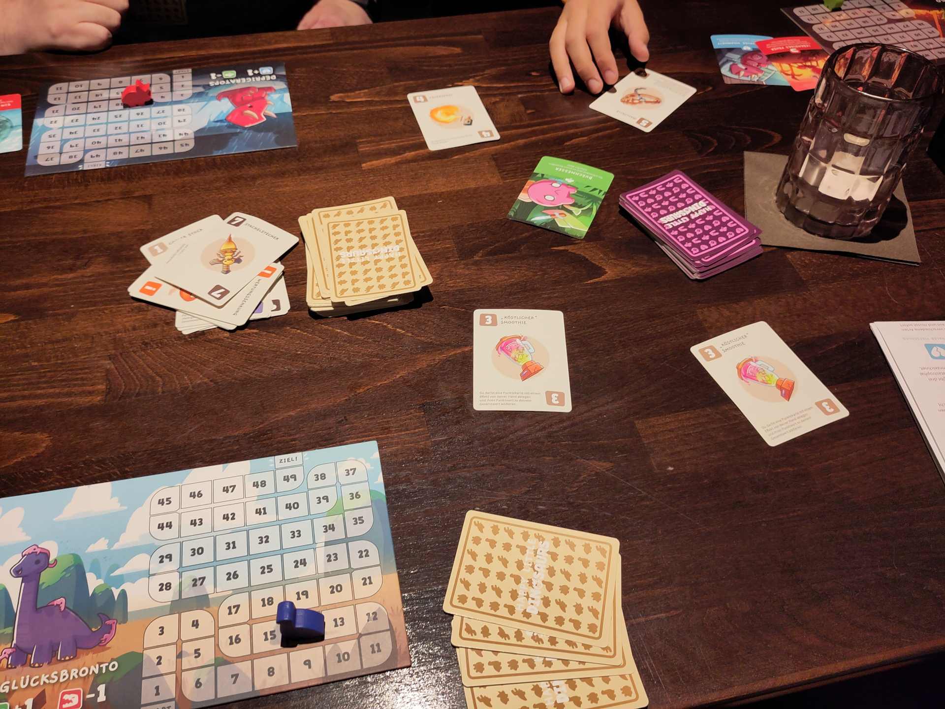 The image shows a dark wooden table with a game of 'Happy little dinosaur' set up. There are three scoreboards for three people, each showing a different dinosaur with a little wooden dinosaur in the matching color indicating the scores. In the middle of the table are two piles of covered cards and some open cards showing unusual weapons like a bee attached to a sword.