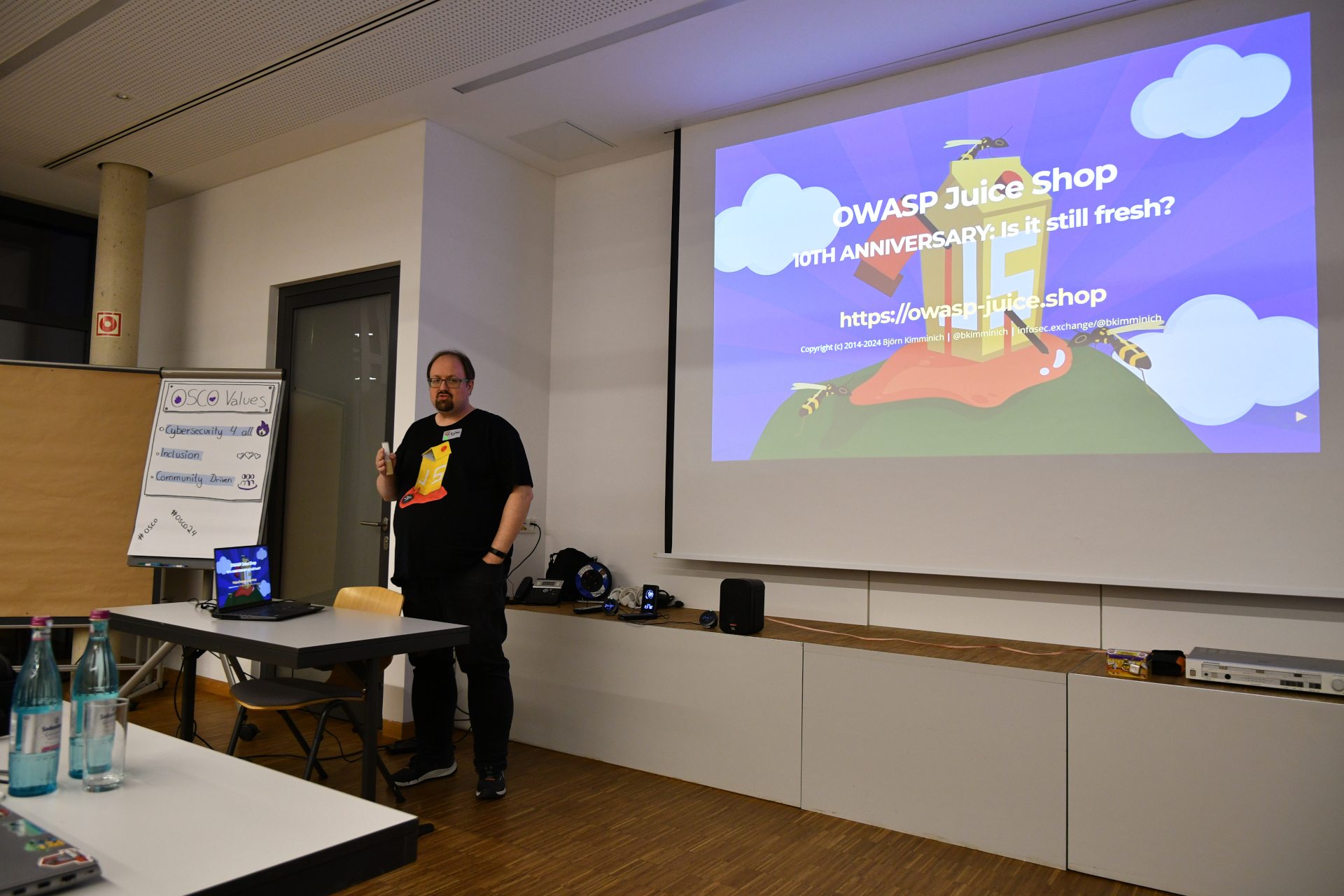 One of the two keynotes: OWASP Juice Shop's 10th Anniversary by Björn. Björn stands in front speaking to the audience behind the photograph. On the right site is the first page of his presentation on the wall. On his left site is the osco values flipchart.