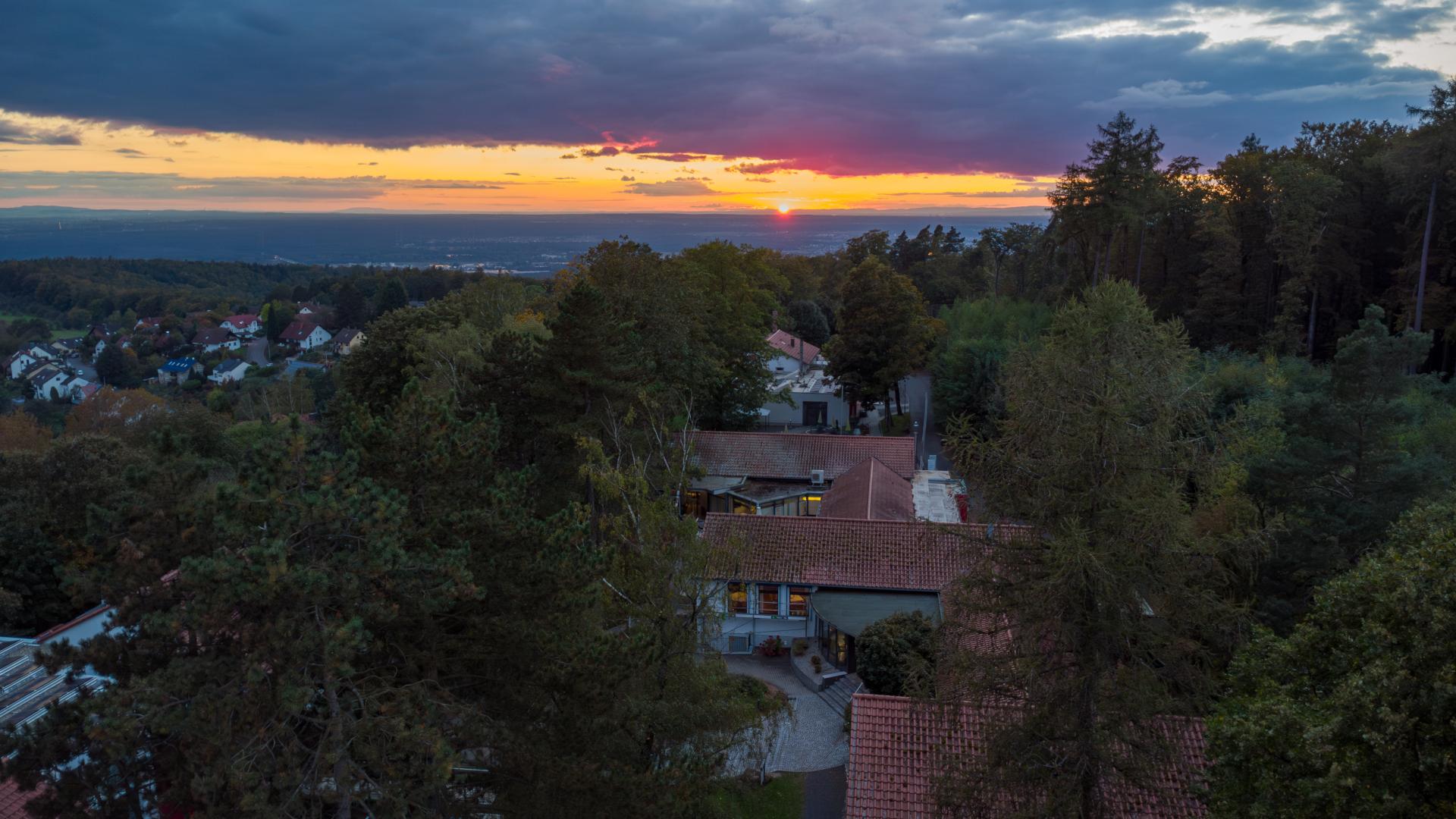 A drone shot of the Seminarzentrum Rückersbach at dusk. The view is from above the building directed at the settling sun at the horizon. The sky is cloudy and they appear very dark above the sun. The Seminarzentrum is surrounded by trees. On the left and in the background are more houses.