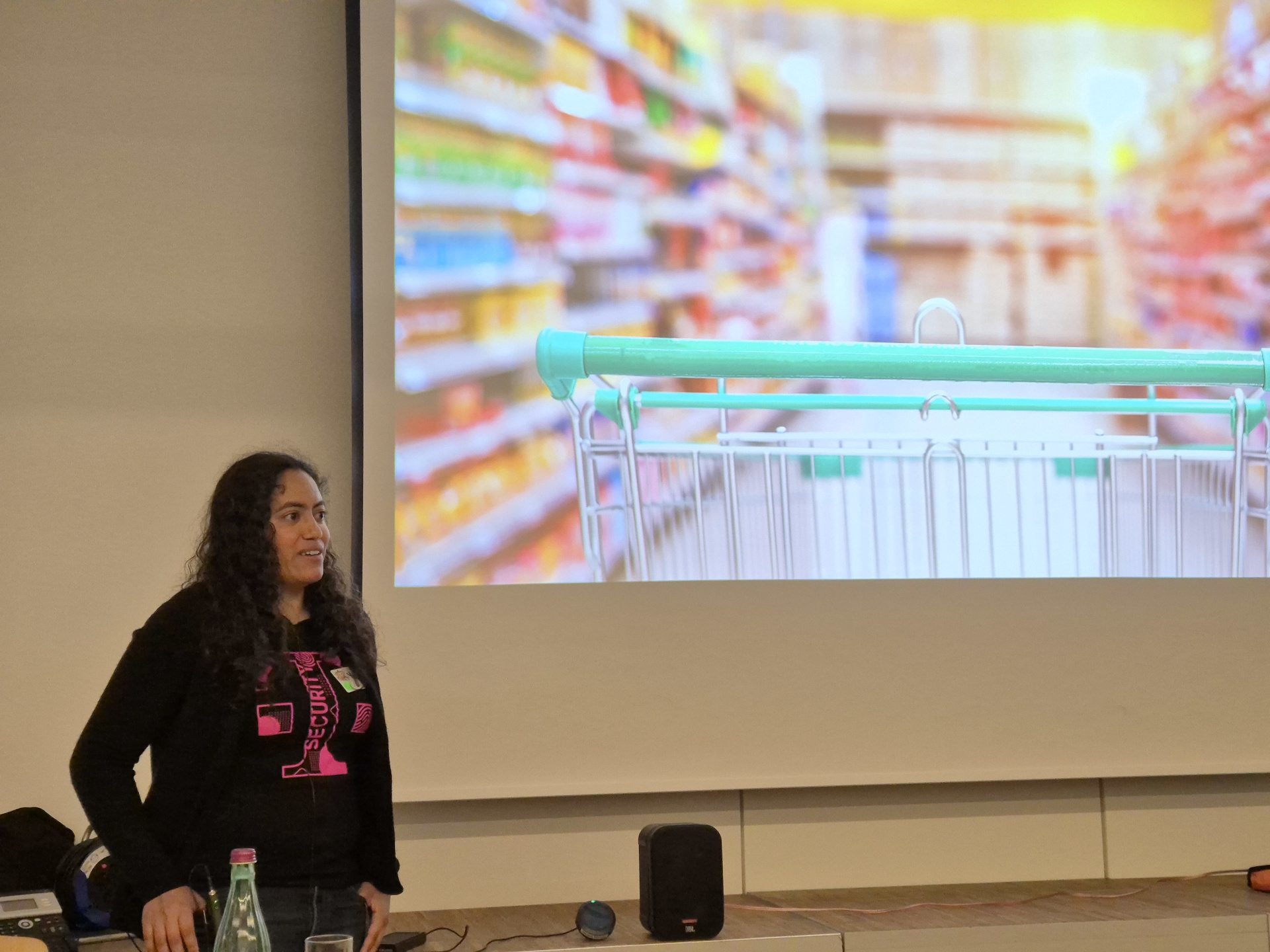One of the two keynotes: Red Teaming by Yvonne. Yvonne stands in the left part of the photo. The most part is a slide of her keynote. A picture of a shopping trolley in a shop.