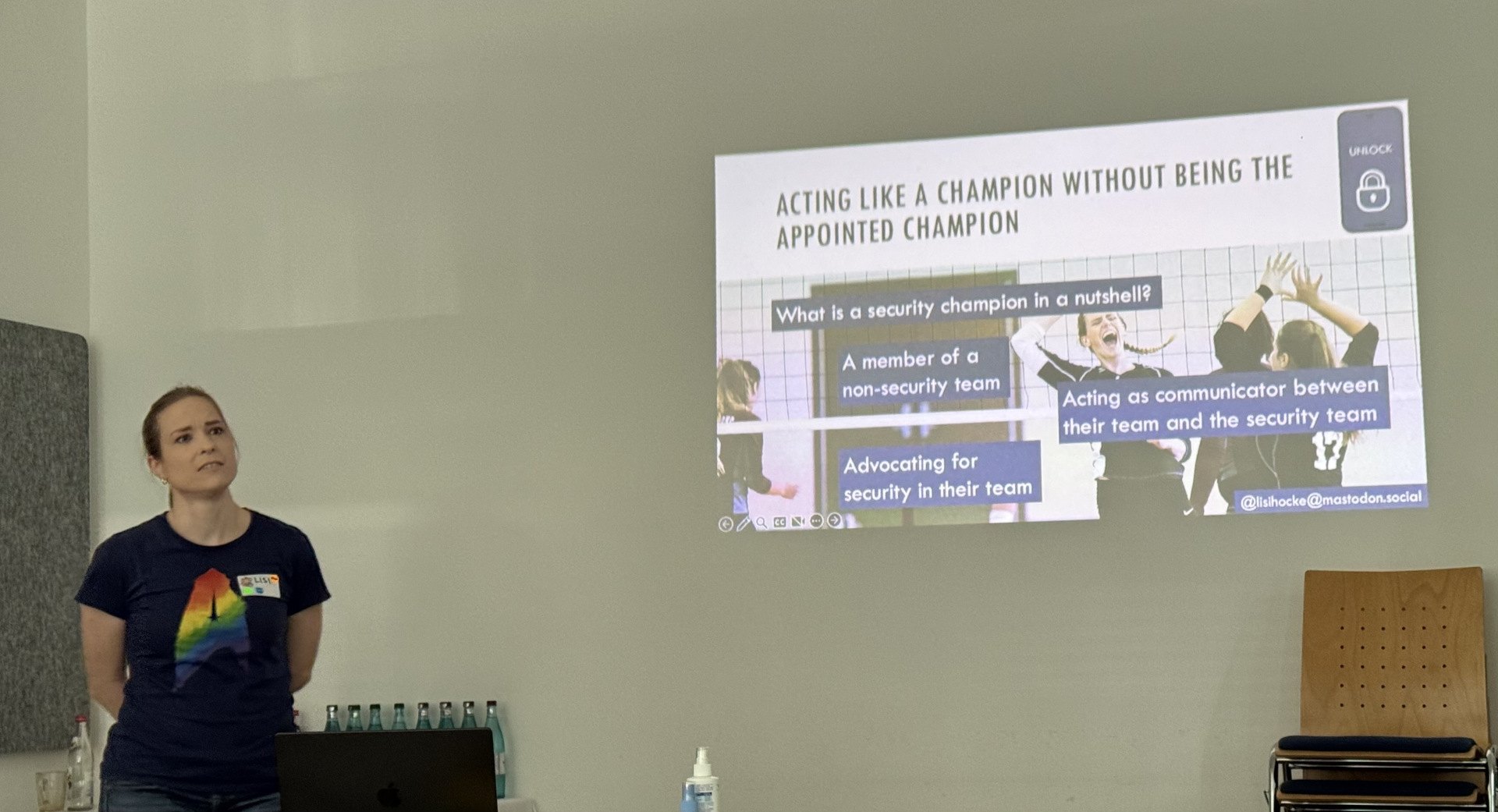 A woman with black tshirt and rainbow colored Star Trek logo stands with her hands behind her back looking to the audience behind the photograph. To her right is her presentation at the wall. The current slide is titled 'Acting like a champion without being the appointed champion'. The slide is about security champions and their definition.