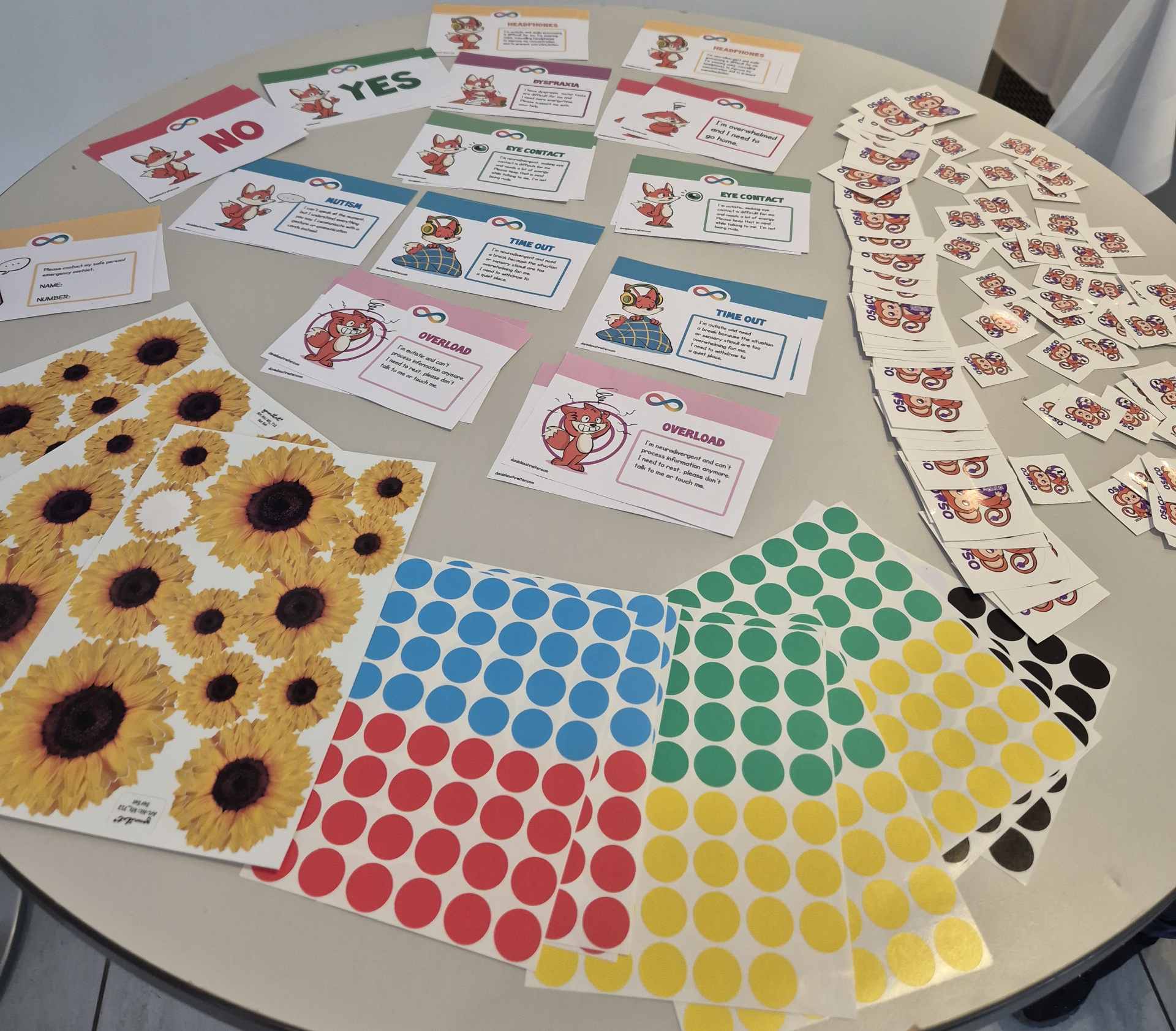 A table with supportive stuff to tell others your needs. Sunflower stickers to show invisible disability. Colored stickers to show if you want to be on photos or not. Cards to show mental overload or that you need a timeout. And some osco 2024 stickers.